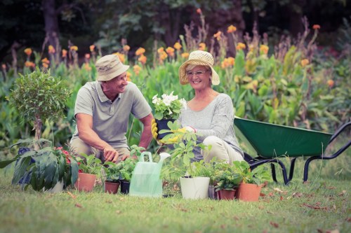 The image depicts two elderly individuals, a man and a woman, sitting together in a well-maintained garden with lush green grass and a variety of potted plants and flowers. The woman is holding a white flowering plant, while the man appears to be engaged in a conversation or examining the plants. Both are wearing sun hats and casual gardening clothing, suggesting outdoor gardening activities on a bright, clear day. Behind them, a vibrant border of purple and orange flowering plants adds colour and natural beauty to the landscaped outdoor space, which appears to be part of a residential garden in Notting Hill. To their left, a small shrub is visible, and a few gardening tools, including a watering can, are placed nearby, indicating ongoing yard maintenance work. A green wheelbarrow is positioned to the woman's right, further emphasizing the gardening context. The scene reflects a tidy, thoughtfully cultivated garden area designed for relaxation and outdoor plant care, aligning with professional gardening services offered by Landscapers Notting Hill in the local area, with natural lighting accentuating the garden's healthy plants and inviting atmosphere.