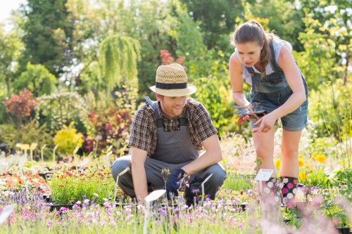 In a vibrant backyard garden during daytime with bright, natural sunlight, two women are engaged in gardening activities. One woman, wearing a wide-brimmed straw hat, a plaid shirt, and dark trousers, is kneeling close to the ground, tending to a bed of colourful flowering plants, including purple, pink, and white blooms. The other woman, dressed in a sleeveless top, denim shorts, and holding garden shears, is leaning forward, assisting with pruning or plant care. Behind them, the garden features a well-maintained lawn with lush, green grass, bordered by neatly arranged flower beds and a mixture of shrubs, bushes, and small trees adding vertical interest. A wooden garden shed is visible in the background, along with mature trees providing shade and a natural backdrop. The scene, set in Notting Hill, showcases a tidy outdoor space designed for gardening and leisure, with a focus on greenery and flowering plants, emphasizing a professional yet inviting environment designed for gardening and outdoor maintenance services.
