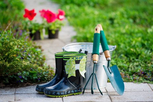 The image depicts a gardening scene set on a paved outdoor space in a garden, with a pair of wellington boots featuring a floral pattern resting on the ground. Next to the boots are gardening tools, including a trowel, a small hand rake, and a pruning shear, all positioned upright. Behind the tools is a clear plastic bucket, possibly for watering or holding garden waste. In the background, there are vibrant pink and red flowers, along with lush green foliage, indicating a well-maintained garden area. The garden surface appears to be made of grey paving stones, and the environment is bright, suggesting daylight with natural light illuminating the scene. The overall setting is indicative of typical outdoor gardening activities, reflecting care and maintenance of a residential garden space, which is characteristic of services offered by companies like Landscapers Notting Hill in the W11 postcode area of London.