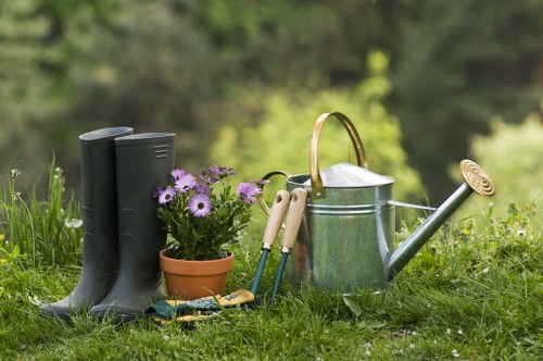 A collection of gardening tools and accessories arranged on a lush green lawn in a garden setting, including a pair of tall black rubber boots positioned to the left, a terracotta pot filled with purple and white flowers, and various small hand tools such as pruning shears and a trowel placed nearby. In the center, there is a silver metal watering can with a curved handle and long spout, alongside a green garden hose connected to it. The background features blurred foliage and trees, indicating an outdoor space in a residential garden environment, with natural daylight illuminating the scene. The arrangement suggests preparations for garden maintenance or planting tasks, aligning with gardening services offered by landscapersnottinghill.co.uk in the Notting Hill area, completing a vivid and detailed visual of outdoor gardening tools in use during mild weather.