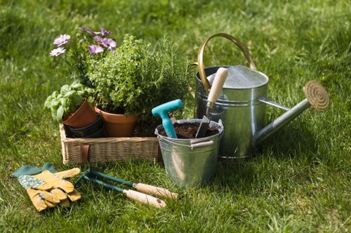 A neatly maintained garden scene featuring a well-kept lawn with lush, green grass occupying most of the foreground. In the middle ground, there is a collection of gardening tools and supplies arranged on the grass, including a pair of yellow gardening gloves, a small blue-handled hand trowel, and a pair of pruning shears. To the left, a rectangular wicker basket holds potted plants, including purple flowering plants, green leafy herbs, and assorted foliage. To the right, a silver watering can with a wooden handle rests on the grass, alongside a plastic bucket containing soil and a smaller garden trowel. The background suggests a typical residential outdoor space, with a uniform green lawn and natural lighting indicative of a clear day, suitable for garden upkeep and landscaping efforts. The scene underscores gardening activities common in property maintenance by Landscapers Notting Hill, aligning with services such as lawn care, planting, and outdoor improvements in the Notting Hill area and postcode W11.