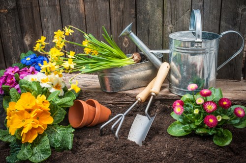 A garden scene featuring a freshly turned soil bed bordered by a weathered wooden fence. In the foreground, vibrant flowering plants including yellow primroses, purple violets, and pink-adonised primulas add colour to the landscaped area. Gardening tools such as a hand rake, trowel, and fork are arranged on the soil, alongside a small terracotta pot and a metal watering can with a handle, indicating recent planting or garden maintenance. To the right, a cluster of potted plants and flower containers sit adjacent to the soil bed, contributing to a well-maintained outdoor space typical of a residential garden in Notting Hill. Natural daylight and mild weather illuminate the scene, highlighting the textures of the soil, plant leaves, and gardening equipment, supporting professional gardening services including lawn care and plant upkeep for local gardens in the area.