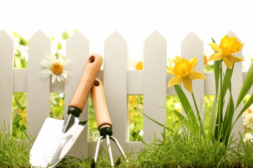 A close-up view of a small garden scene in a residential outdoor space, featuring a white picket fence with vertical slats in the background, partially obscured by bright yellow daffodil flowers and white daisies with yellow centers growing beside it. In the foreground, there is lush, dense green grass with a slightly uneven surface, alongside a set of gardening tools including a trowel and a cultivator with wooden handles, resting on the grass. The background suggests a sunny day with natural lighting illuminating the garden, highlighting the vibrant colours of the flowers and the textures of the grass and soil. This scene reflects typical gardening and outdoor maintenance activities, aligning with professional services offered by landscapers in the Notting Hill area, and showcases elements commonly found in a well-maintained garden space ready for landscaping or general outdoor care.