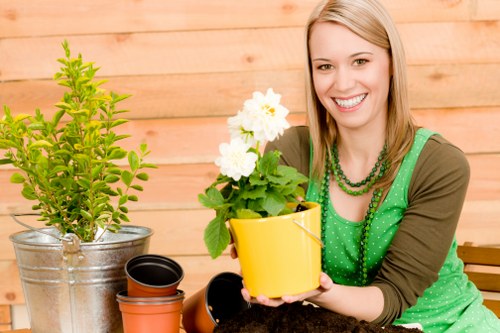 The image shows a smiling young woman with long blonde hair sitting outdoors in a garden setting, holding a small yellow flower pot containing a flowering plant with white blossoms. In front of her on a table are several empty pots of varying sizes, including a black plastic one, a terracotta-colored one, and a smaller black one, suggesting she is engaged in gardening activities. To her left, there is a large green shrub planted in a metallic container, and the background features a natural wooden fence or wall, indicating a well-maintained private garden or outdoor space typical of Notting Hill properties. The scene is well-lit with natural daylight, giving a warm and inviting atmosphere suitable for gardening or landscaping services offered by professionals like Landscapers Notting Hill. The arrangement and details highlight typical elements of outdoor garden care, planting, and container gardening, reflecting a neat, cultivated environment in an urban green space.