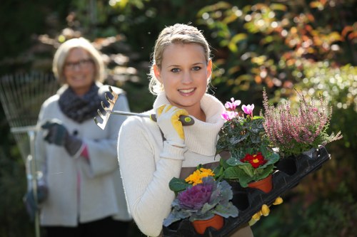 A young woman with light blonde hair pulled back, smiling at the camera while holding a black tray filled with various flowering plants and small shrubs, in a garden surrounded by lush greenery, including trees and shrubs with red and green foliage. She is wearing a white sweater and yellow gardening gloves, indicating outdoor gardening activity. In the background, an older woman with glasses and short blonde hair is slightly blurred, also dressed in gardening attire, holding a garden rake, suggesting a collaborative gardening effort in a well-maintained landscape area. The garden features a manicured lawn, flower beds with colorful blooms, and natural lighting that highlights the vibrant plant colors and outdoor setting typical of a professional garden maintenance scene in Notting Hill, London, fitting with landscaping and gardening services supplied by Landscapers Notting Hill.